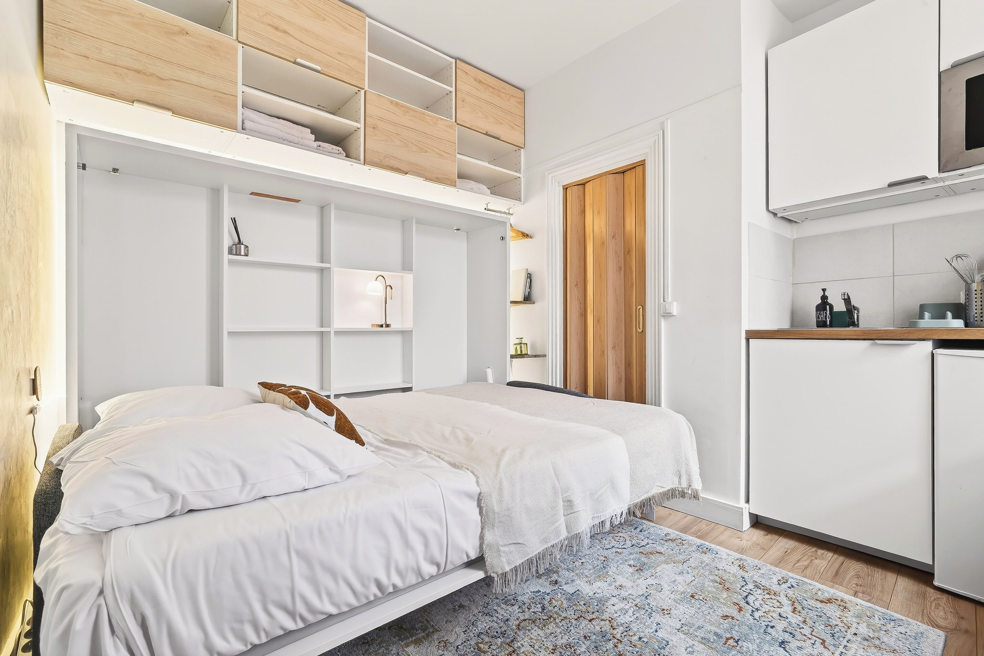 A bedroom with a bed, a built-in wardrobe, a kitchenette, and a door leading to another room.