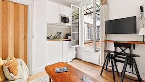 A compact kitchen with a microwave, a small countertop, and a window. There is a wooden table with a book, a sofa with cushions, and a TV mounted on the wall. - Lovely Studio 10 mn walk to Louvre & Latin Quarter (Paris)