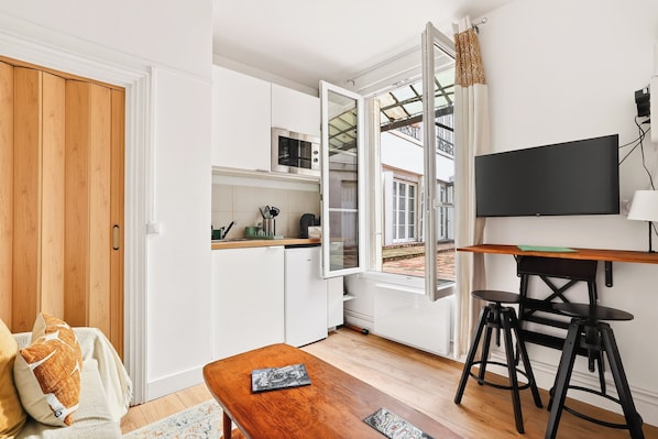 A compact kitchen with a microwave, a small countertop, and a window. There is a wooden table with a book, a sofa with cushions, and a TV mounted on the wall. - Lovely Studio 10 mn walk to Louvre & Latin Quarter (Paris)