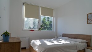Een slaapkamer met een bed, nachtkastje, raam met uitzicht op groen en een aan de muur hangend schilderij.
