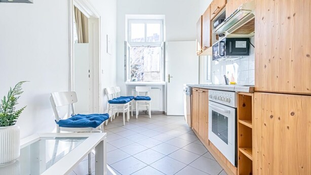 A kitchen with wooden cabinets, a white countertop, and a window. There are two white chairs with blue cushions. - Nice Room for Longstays @Margareten, Up to 2P (Vienna)