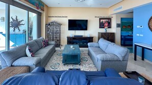 A living room with two grey sofas, a coffee table, a TV, and a bookshelf. - Waves Relaxation Oceanfront 2BR Family Condo (Puerto Peñasco)