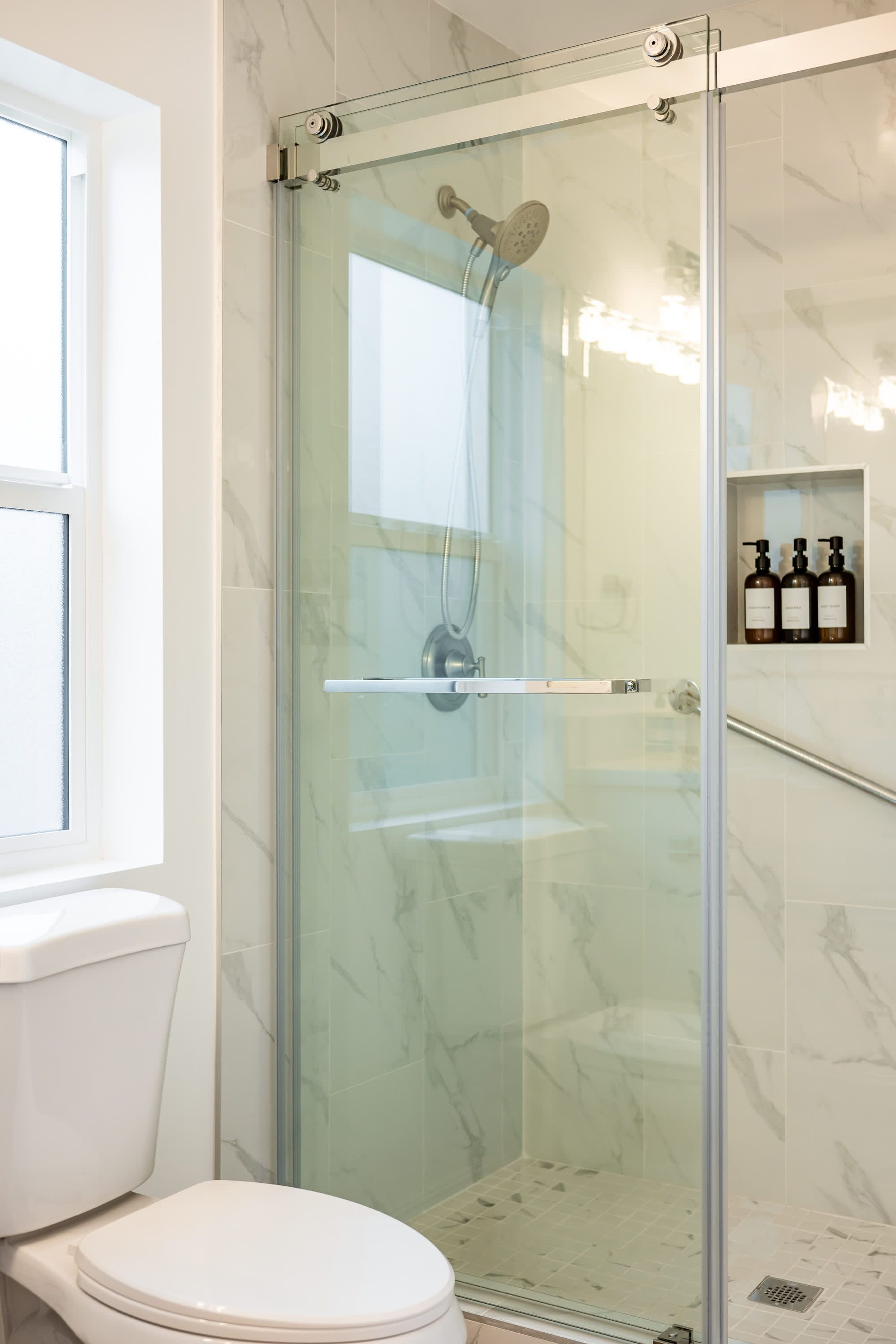 A glass-enclosed shower with a handheld showerhead, a toilet, and a shelf with toiletries.