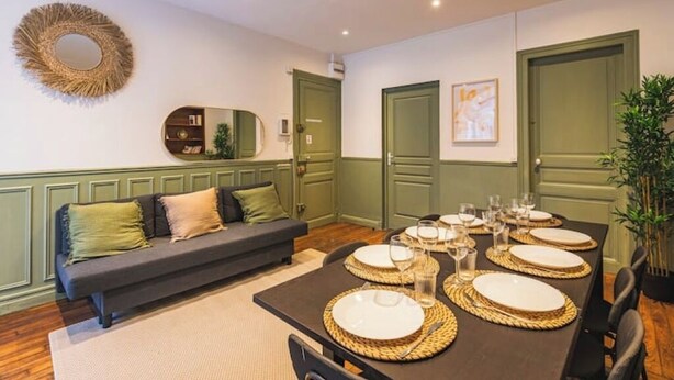 A dining area with a long table set for six, surrounded by chairs. There is a sofa with cushions against the wall, and a mirror above it.