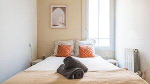 A neatly made bed with a rolled towel, two pillows with a geometric pattern, a framed picture on the wall, and a window with natural light.
