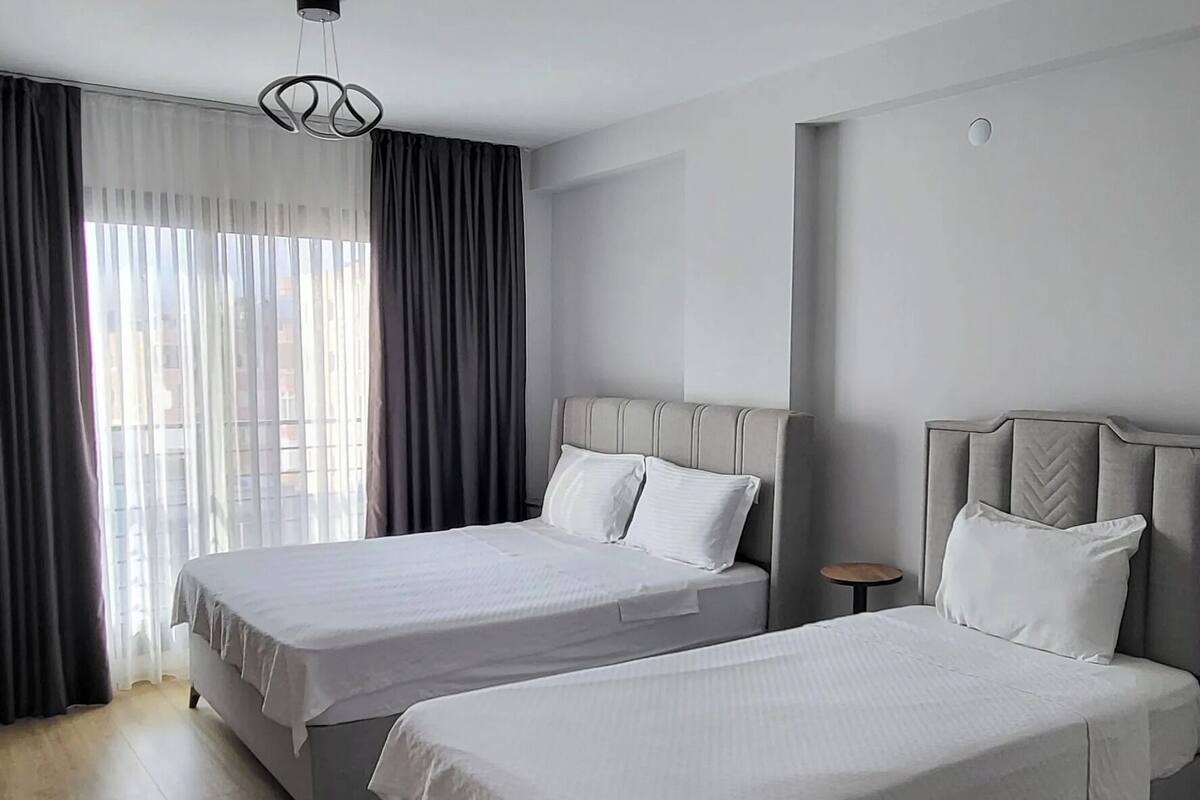 Habitación de hotel moderna con dos camas, un gran ventanal con cortinas, una lámpara de araña contemporánea y suelo de madera.