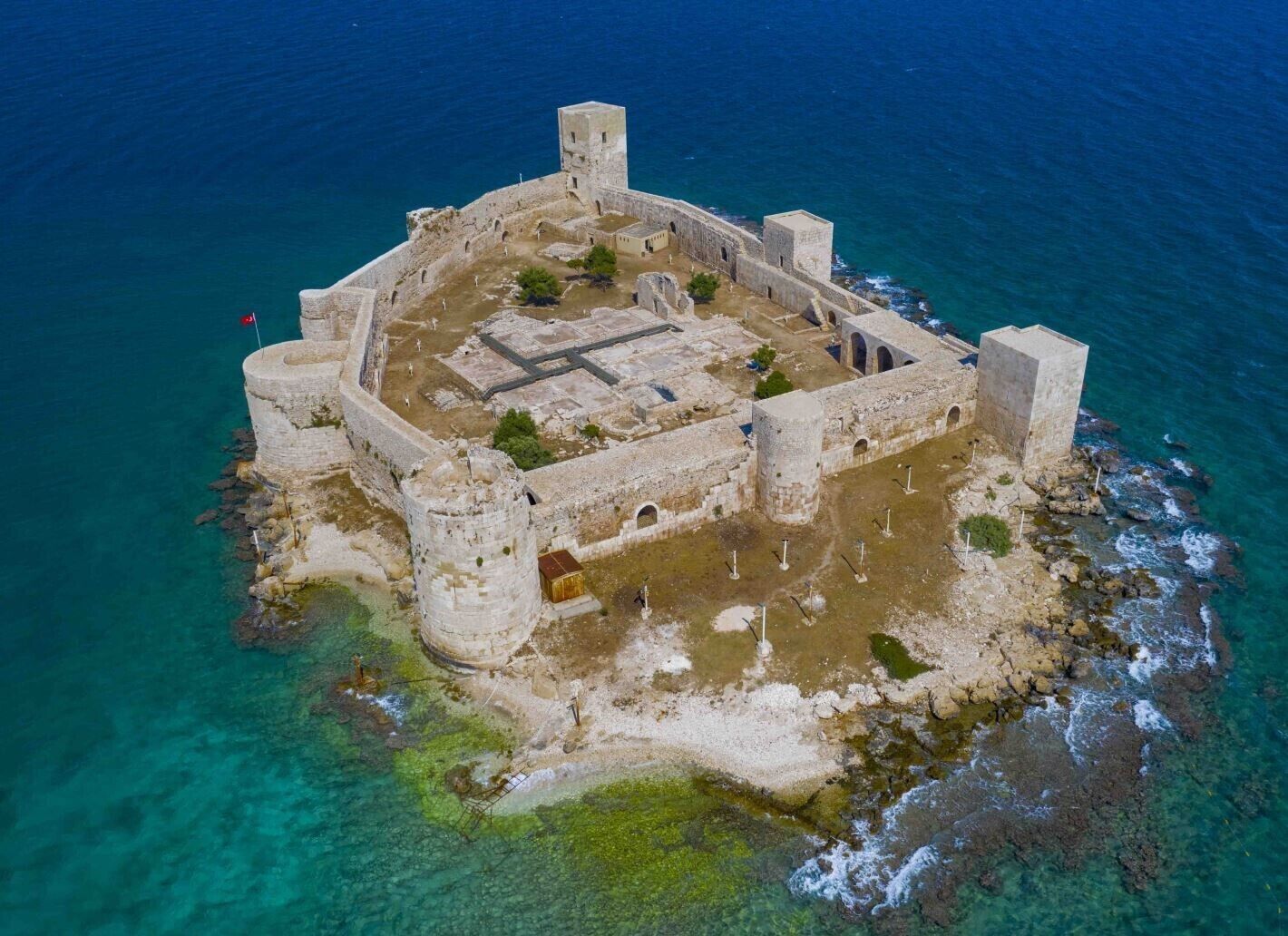 Vista aérea de una fortificación histórica con un patio central, rodeada de muros y torres, situada en una isla rocosa con aguas azul turquesa.
