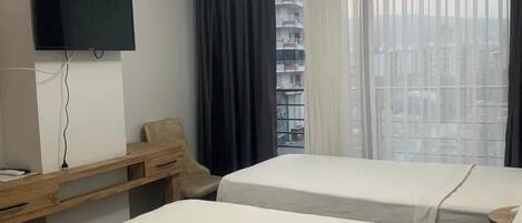 Hotellihuone, jossa on kaksi sänkyä, työpöytä, tuoli, televisio ja näkymä kaupunkiin ohuiden verhojen läpi.