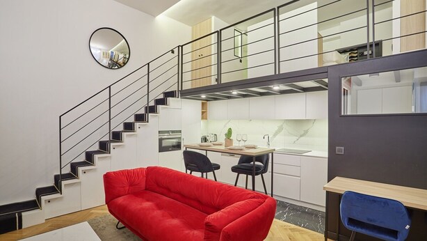 A modern kitchen with white cabinets, a dining area with two chairs, a red sofa, and a staircase with a railing. - Duplex Center of Paris (Paris)