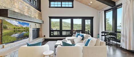 A spacious living room with a stone fireplace, large windows, and a view of the outdoors.