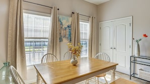 Un coin repas avec une table en bois, des chaises blanches, un vase de fleurs et une vue sur l’extérieur grâce à de grandes fenêtres ornées de rideaux.