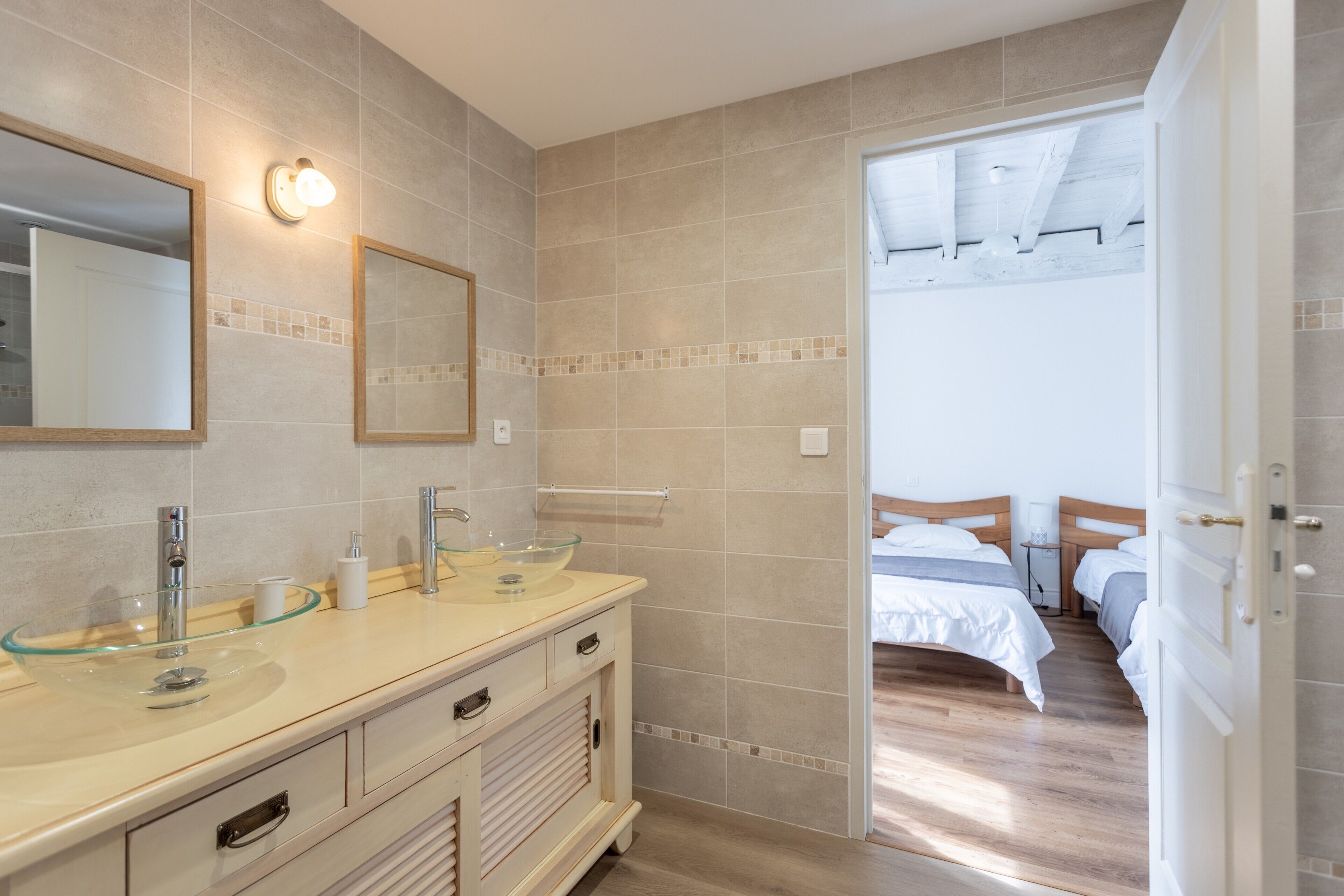Baño con dos lavamanos, un lavabo de cristal y un espejo. Hay una puerta que da a un dormitorio con dos camas.