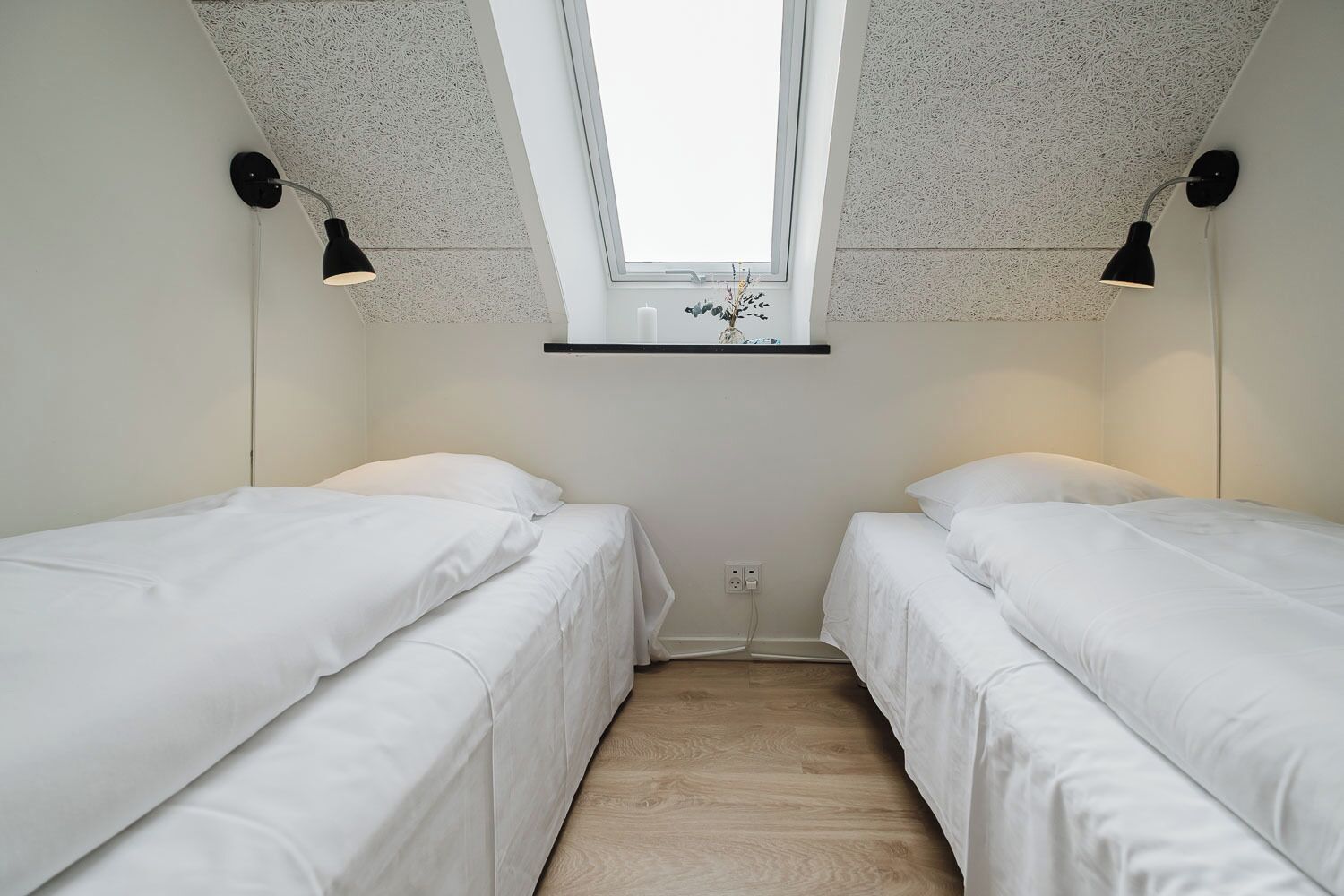 Una habitación con dos camas individuales, un tragaluz y un pequeño estante sobre la ventana.
