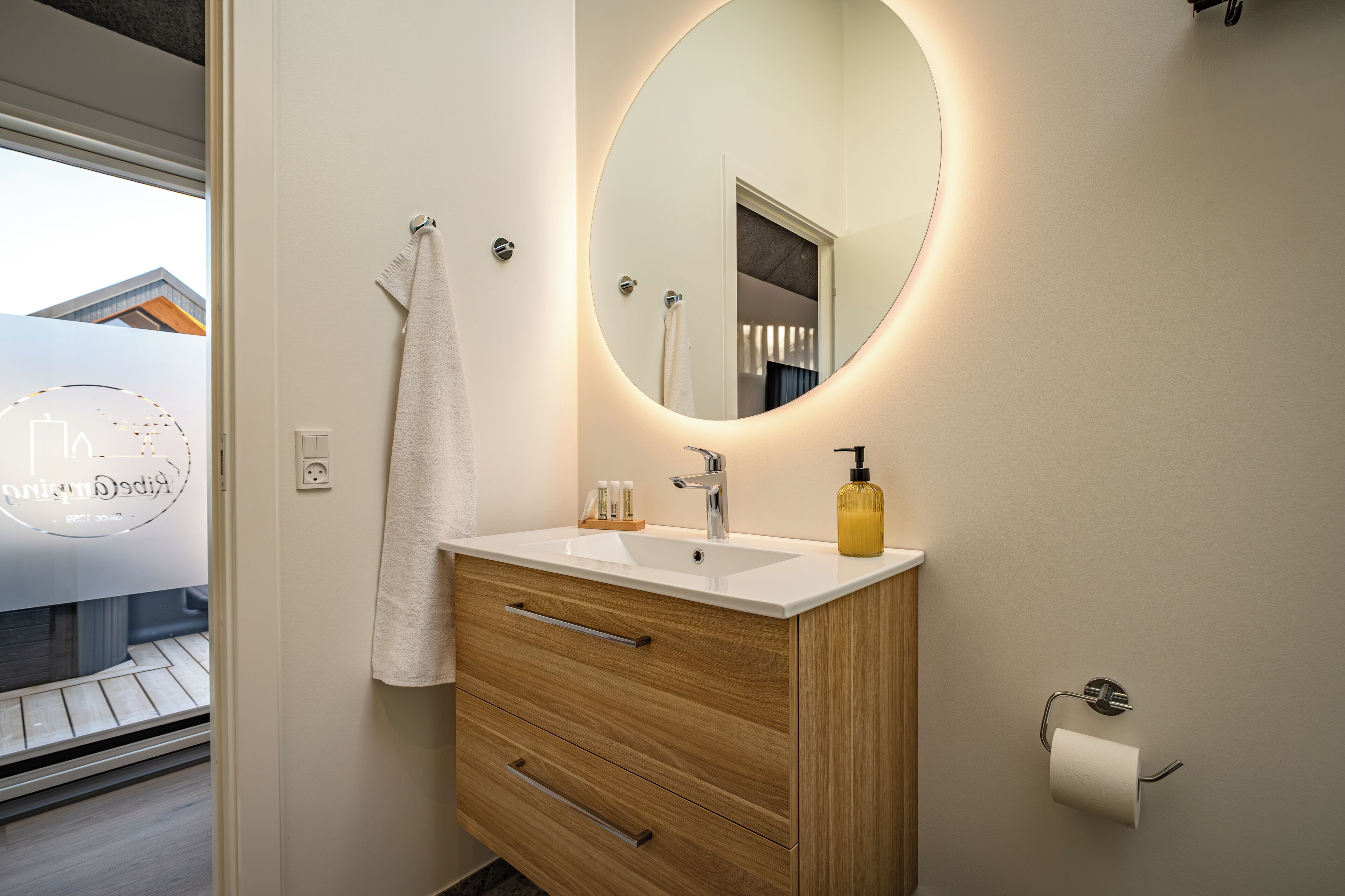Een moderne badkamer met een witte wastafel, een houten badkamerkastje, een ronde spiegel en een toiletrolhouder.