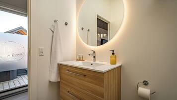 Een moderne badkamer met een witte wastafel, een houten badkamerkastje, een ronde spiegel en een toiletrolhouder.
