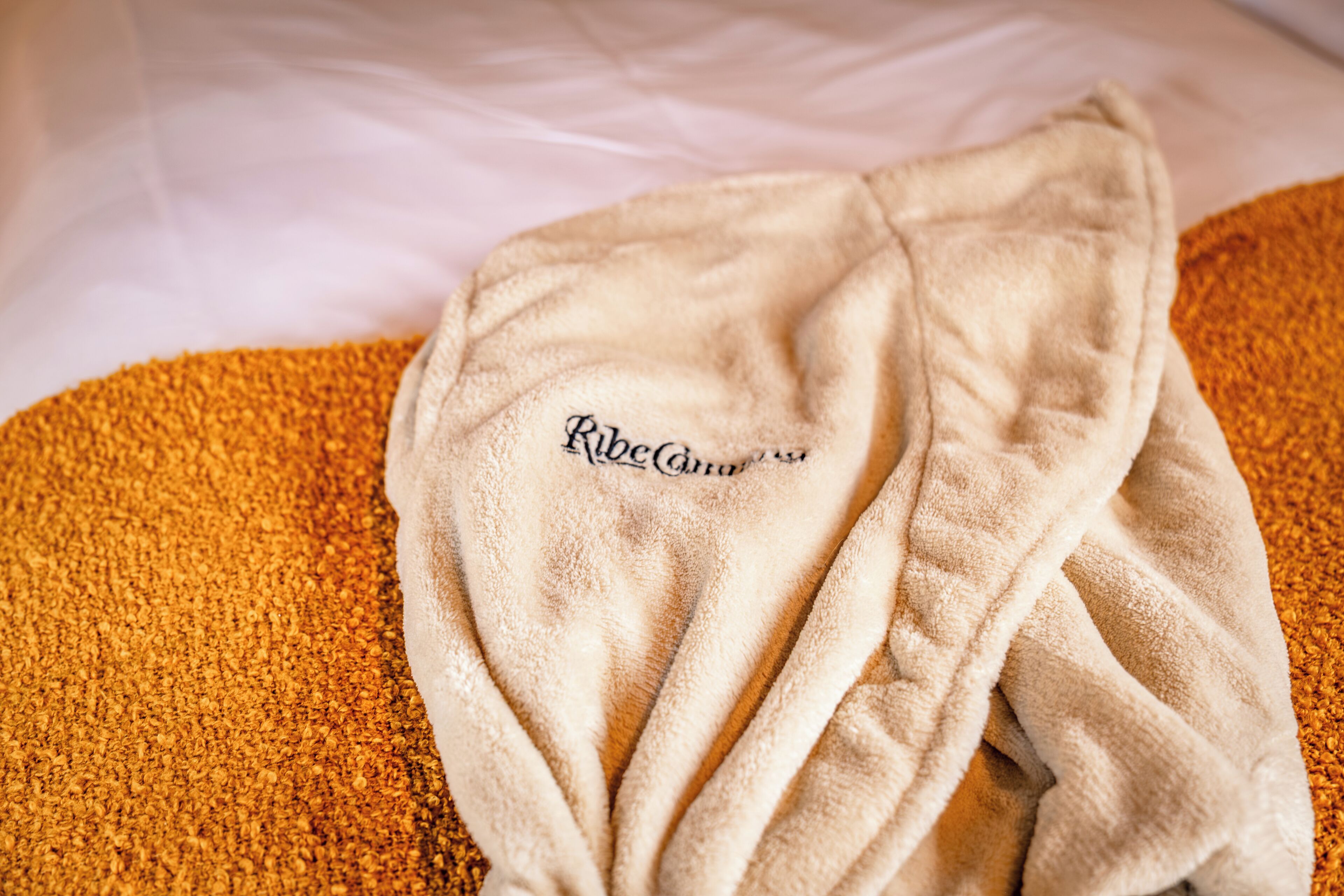Un albornoz beige con el bordado 'Ribeauville', sobre una cama con un edredón anaranjado texturizado.