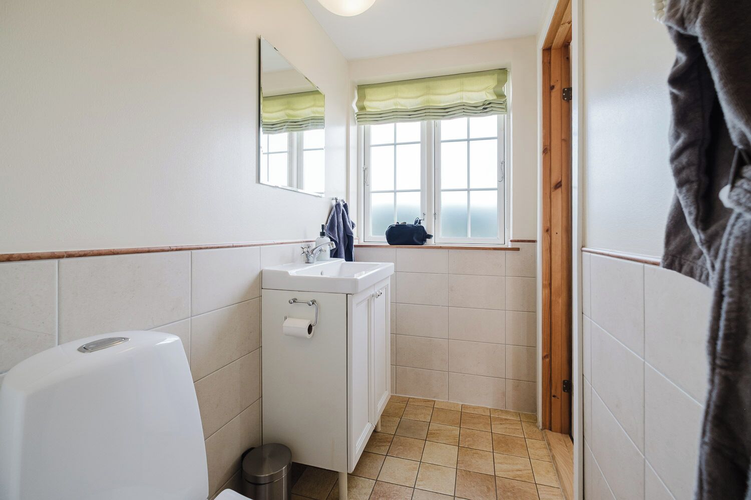 A small bathroom with a toilet, sink, and mirror. There is a window with green valance and a towel hanging on the wall.