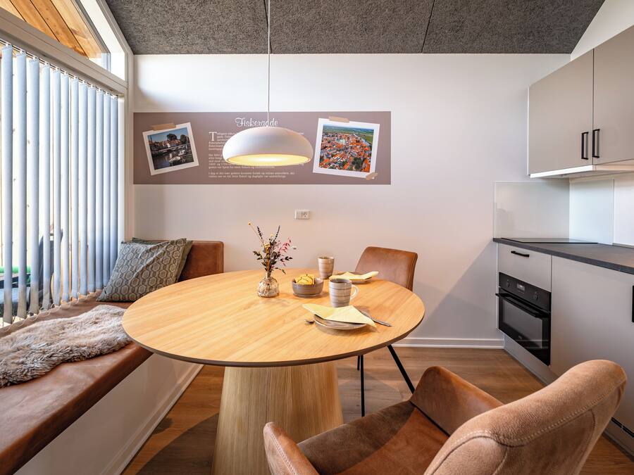Una cocina moderna con una mesa redonda de madera, asientos y vistas de la ciudad a través de las persianas.
