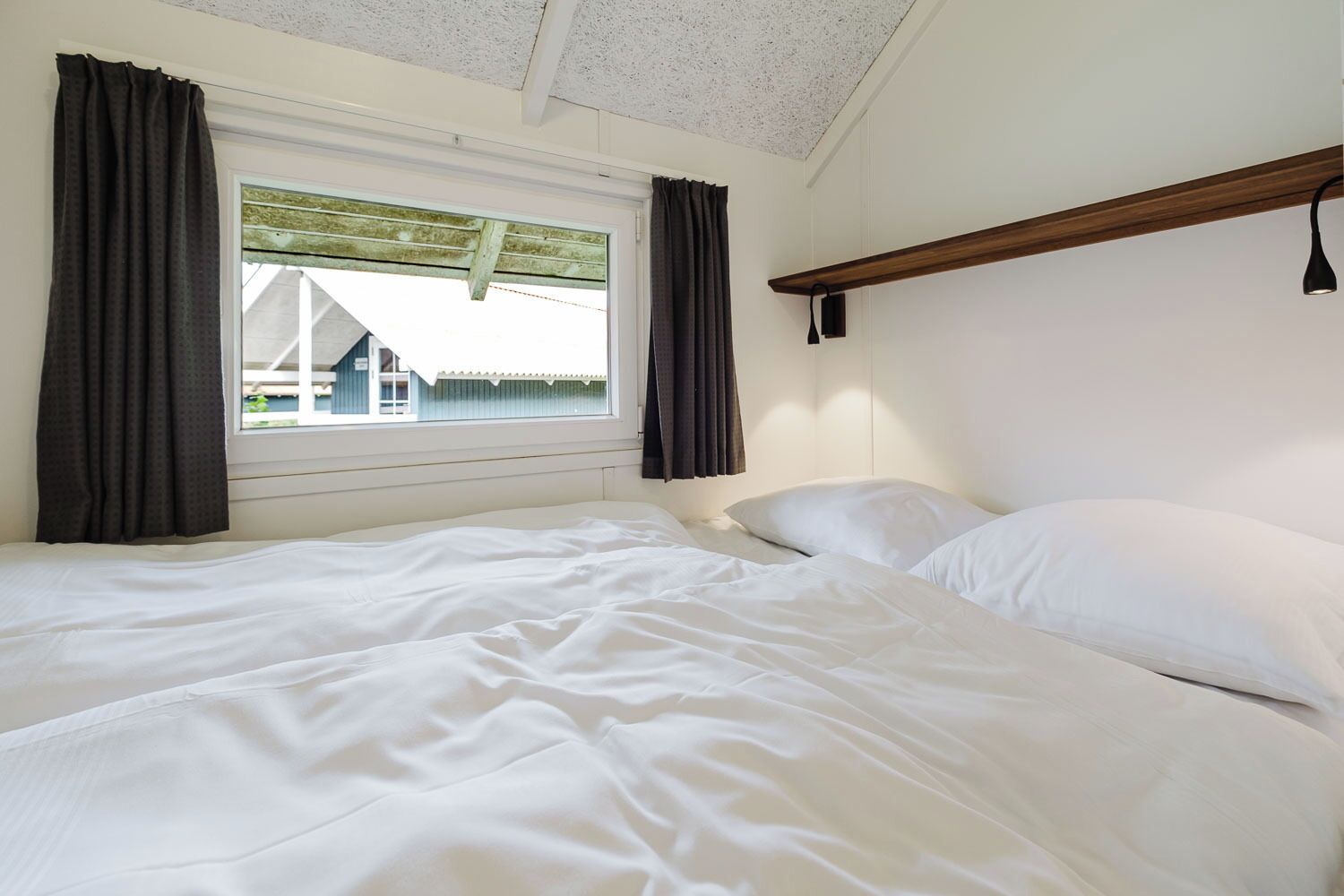 Ein Schlafzimmer mit einem Bett, zwei Kissen, einem Fenster mit Vorhängen und einer Holzablage.
