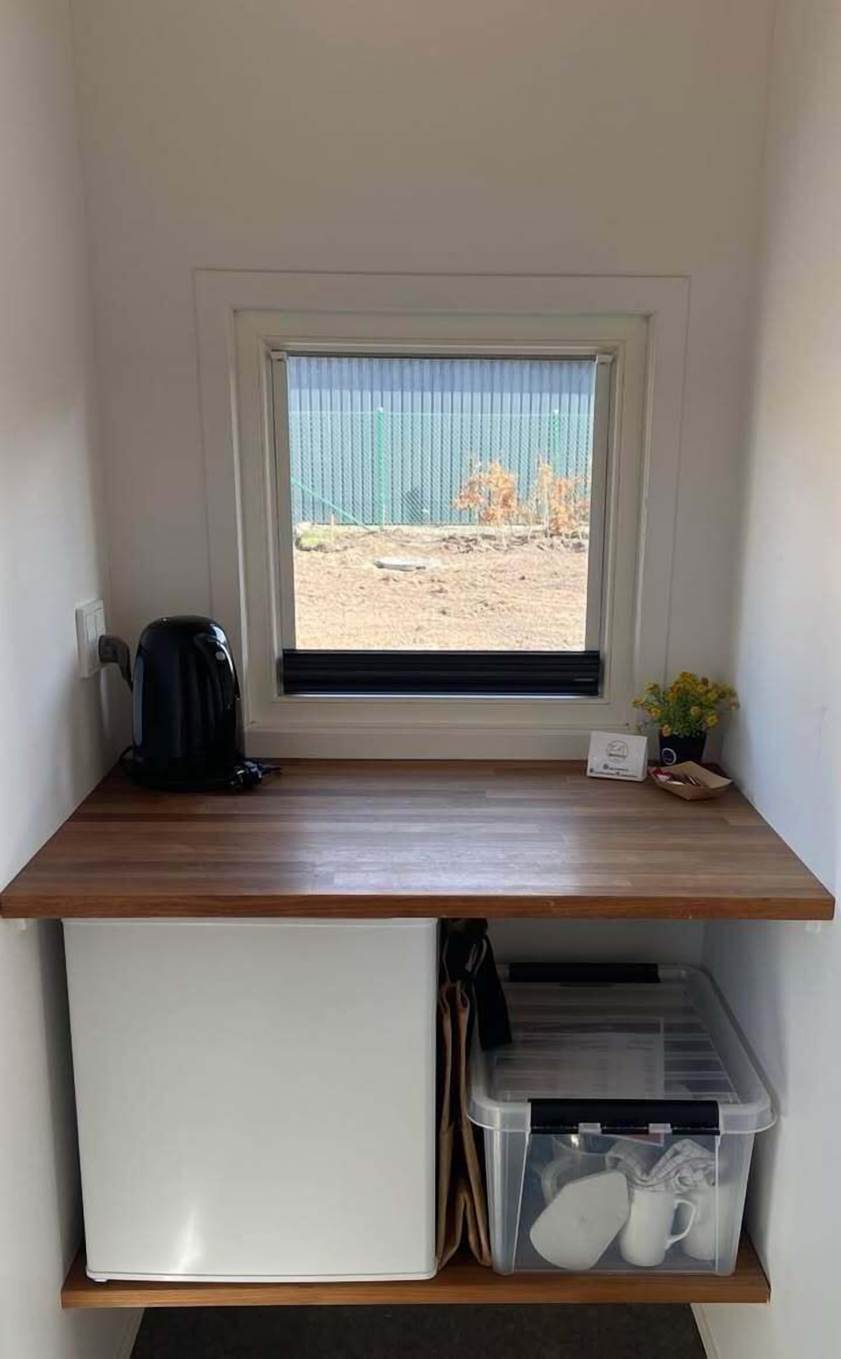 A compact room with a wooden desk, a small refrigerator, a window with a view of a fenced area, and a potted plant.