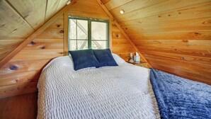 A cozy attic bedroom with a wooden sloped ceiling, a large bed with a white textured bedspread and two pillows, and a window with a green frame. - Cozy Couples Retreat w/ Porch on Bayou Teche! (New Iberia)