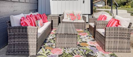 Un patio con muebles de mimbre, un tapete floral y vistas a una casa y árboles.