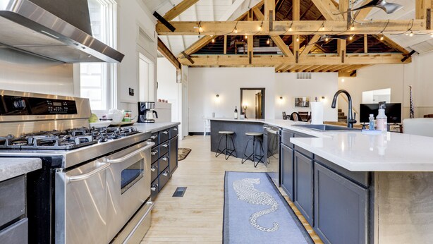A spacious kitchen with high ceilings, wooden beams, a large island, stainless steel appliances, and a cat-themed rug. - 'the Townhall' Northwoods Retreat w/ Large Patio! (Conover)