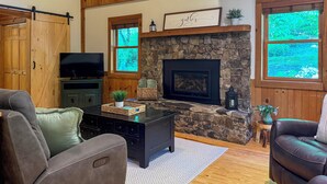 A cozy living room with a stone fireplace, wooden beams, a television, and leather sofas. - Hot Tub, Fish, Ski: Outdoorsy Retreat in Boone! (Vilas)