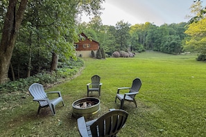 A wooden cabin with a fire pit and Adirondack chairs in a grassy yard.