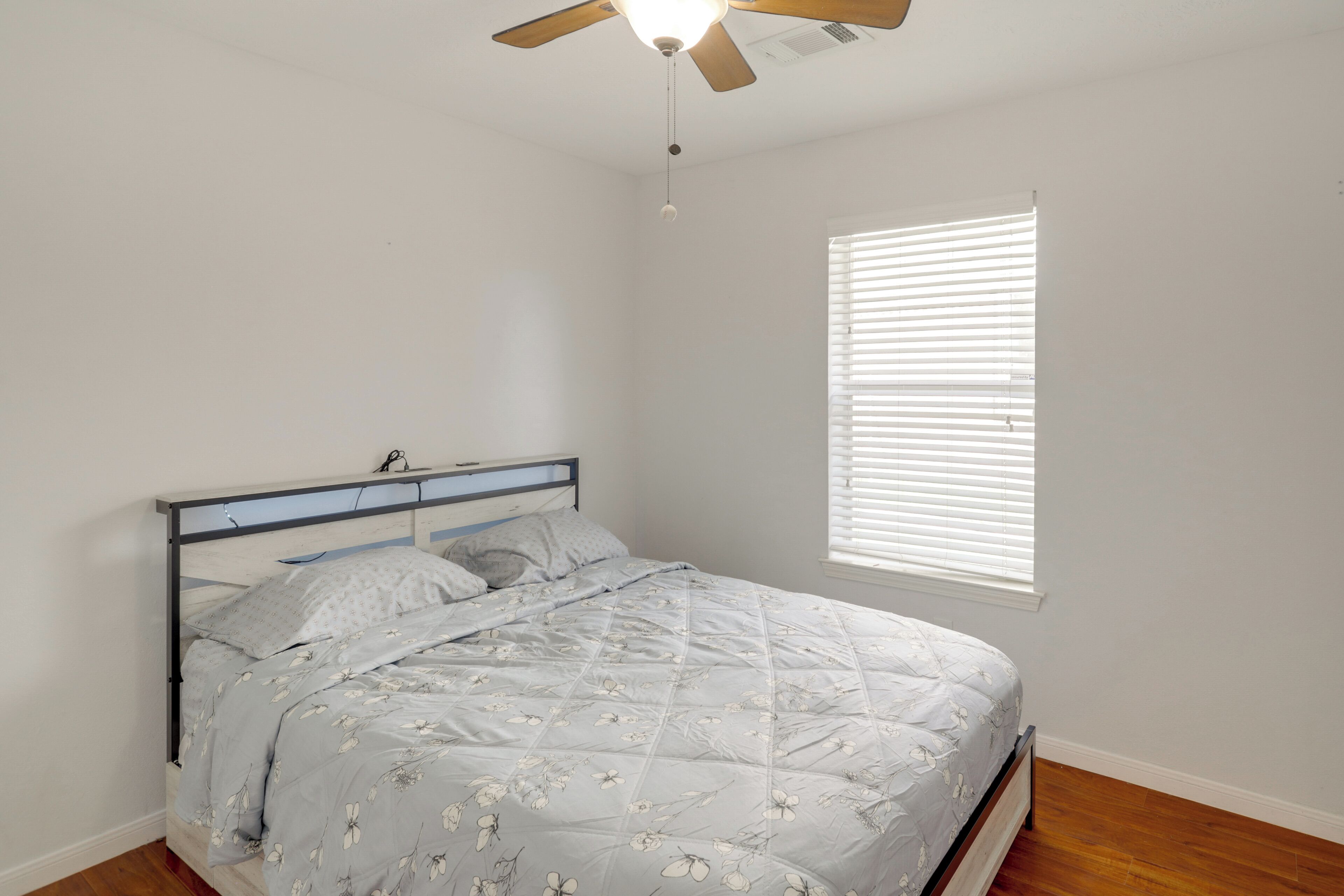 A bedroom with a bed, a ceiling fan, a window with blinds, and wooden flooring.