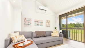 Una sala de estar moderna con un sofá seccional gris, una bandeja de madera con un plato y una taza, y una alfombra blanca. Se observan tres obras de arte en la pared, una ventana con vista a follaje, y una unidad de aire acondicionado en el techo.