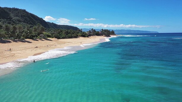 Una playa con agua turquesa cristalina, orilla arenosa y un promontorio boscoso.