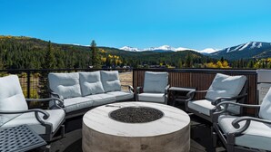 Una terraza con sillas blancas acolchadas y una mesa de piedra circular con un brasero, con vistas a un paisaje montañoso.