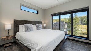 Un dormitorio con una cama grande, un ventilador de techo, una ventana con vistas a árboles y una mesita de noche con una lámpara.