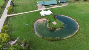 An aerial view of a property with a pond, a white building with a green roof, and a road nearby. - Gather Here: 3,000 Sq Ft Lodge in Michigan's U.P. – Pond, Fire, Sleeps 14 (Wilson)