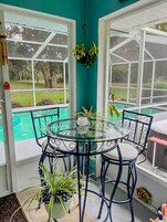 Una mesa con superficie de vidrio y sillas, rodeada de plantas en macetas, en una habitación con paredes verde azulado y ventanas amplias con vista a una piscina.