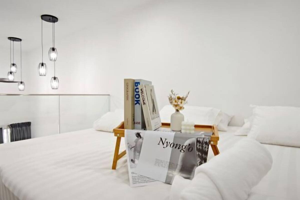 A modern hotel room with a neatly made bed, a bedside table with books and a vase, and pendant lights above.