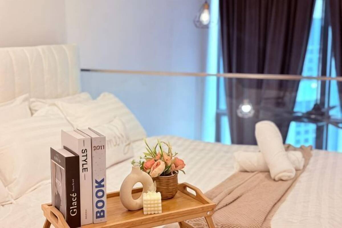 A neatly made bed with pillows, a bedside table with books, a tray with a vase of flowers, and a window with a view.