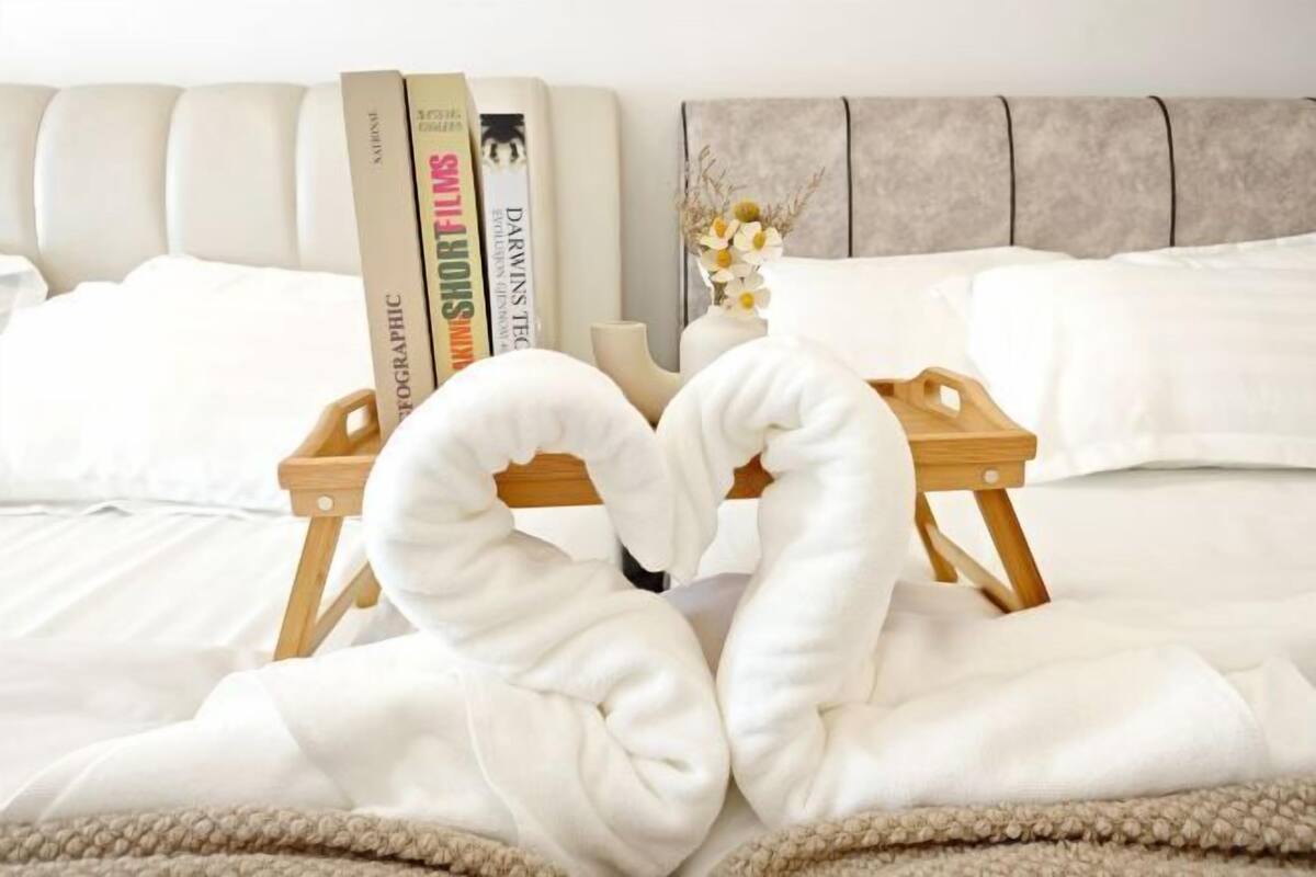 A neatly made bed with a heart-shaped towel arrangement, a wooden tray, and books on a headboard.