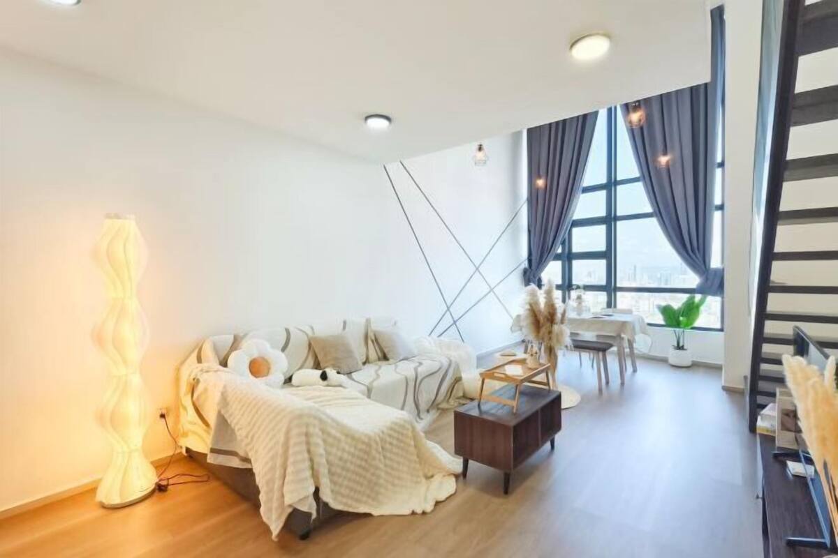 A modern living room with a sofa, a coffee table, and a lamp. There is a window with curtains and a view of the city.