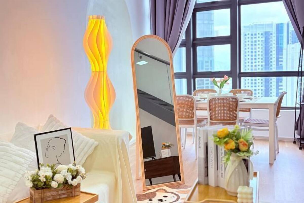 A modern hotel room with a large window, a dining area, and a mirror reflecting the room.