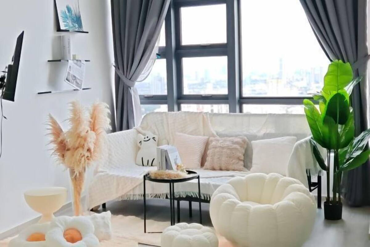 A modern living room with a white sofa, a round white pouf, a large green plant, and a city view through large windows.