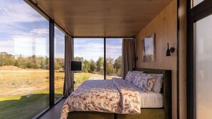 A bedroom with a large bed, a TV, and a view of a grassy field through large windows. - RT Creekside Glass Retreat | Cowboy Pool/Hot Tub (Round Top)