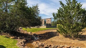 A modern building with a glass facade, surrounded by a natural landscape with trees and a small stream. - RT Creekside Glass Retreat | Cowboy Pool/Hot Tub (Round Top)