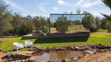 A modern outdoor living area with a glass enclosure, a small pond, and two white chairs.