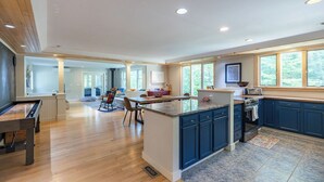 A spacious kitchen with a central island, wooden flooring, and large windows offering a view of the outdoors.