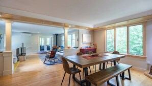 A spacious living area with a wooden dining table, comfortable seating, and large windows offering a view of the outdoors.
