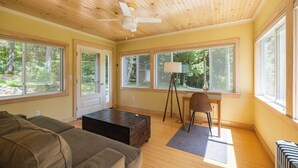 A spacious living room with a wooden ceiling, large windows, a ceiling fan, a desk with a lamp, and a cozy seating area.