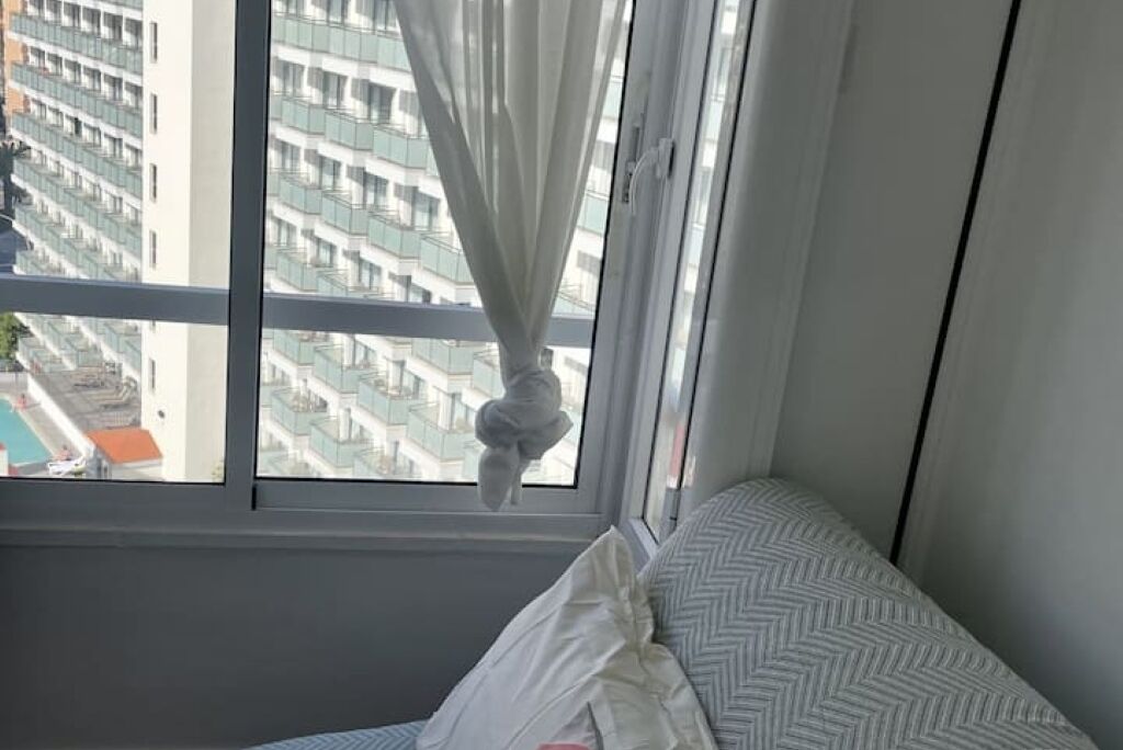 A bedroom with a window, a bed with a patterned bedspread, and a view of a cityscape.