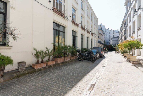 A cobblestone street lined with buildings, parked scooters, and potted plants.